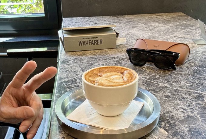 A hand makes a peace sign next to a cappuccino with latte art on a marble table, alongside a Ray-Ban sunglasses case labeled "WAVFARER" and the text "ULTRA COLLECTION" visible on its side, complementing the post's excitement about receiving new Meta glasses.