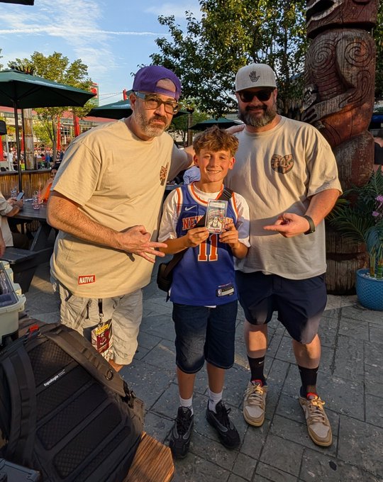 Two men and a young boy posing together outdoors at what appears to be a zoo or park, with the boy holding a small framed item and the men pointing at it.