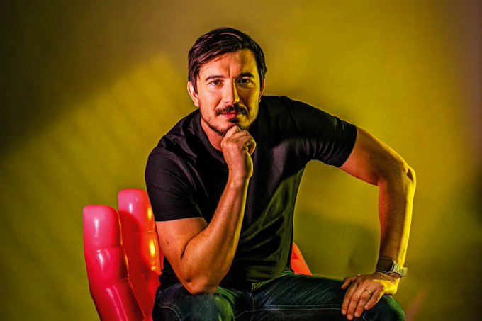 A man with dark hair and a mustache wearing a black t-shirt sits on a red chair against a yellow-green backdrop, resting his chin on his hand.