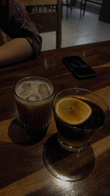 Two iced beverages sitting on a wooden table—one a milky coffee with ice cubes, the other a dark drink garnished with a lemon slice—next to a black smartphone.