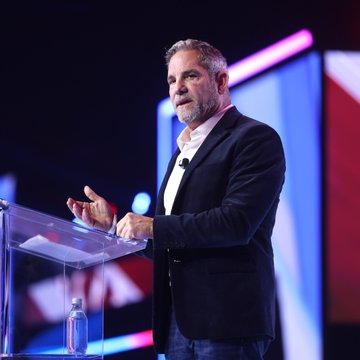 Grant Cardone speaks at a podium during what appears to be a business or motivational event, gesturing with his hand while addressing an audience. The image complements the post about Cardone Capital's recent Bitcoin acquisition, showing Cardone himself as the figure behind the investment announcement.
