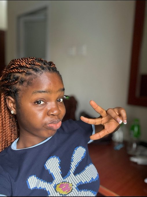 A young woman with reddish-brown braided hair makes a playful face and gestures toward the camera while wearing a navy blue graphic t-shirt.