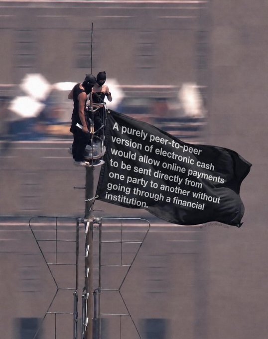 A person atop a high pole or structure holds a large black banner quoting the Bitcoin whitepaper: 'A purely peer-to-peer version of electronic cash would allow online payments to be sent directly from one party to another without going through a financial institution.'