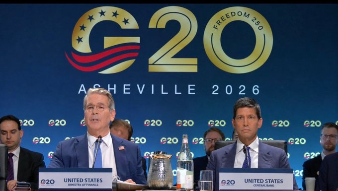 Two U.S. delegates—a representative from the Ministry of Finance and one from the Central Bank, both seated at a "UNITED STATES" placard—attend the G20 summit in Asheville (2026), whose backdrop reads "G20 FREEDOM 250," illustrating the very intergovernmental forum now engaging in dialogue around crypto regulation referenced in the post.