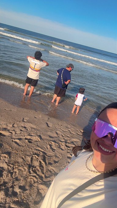 The image shows a group of people standing at the edge of the water, with some looking out into the ocean while others are playing in the shallow water. There are four individuals visible in the scene - two men and two children. The man on the left is wearing a white shirt and black shorts, while the other man is dressed in blue. The child on the right is wearing a pink shirt and sunglasses. A woman with purple sunglasses stands next to them, smiling at the camera.