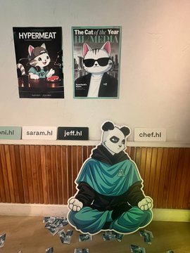 A panda dressed in samurai-like attire sits on the floor in front of a wall displaying posters titled 'HYPERMEAT' and 'The Cat of the Year is MENEKI,' with labels reading 'ni.ni', 'saram.hl', 'jeff.hl', and 'chef.hl' above it.