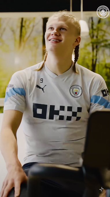 Erling Haaland, wearing his Manchester City #9 jersey featuring the OKX sponsor logo, smiles while looking upward as he relaxes in a chair.