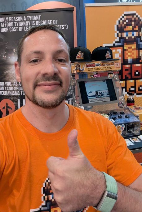 A man in an orange Hero of Bitcoin t-shirt gives a thumbs-up while standing in front of a branded Hero of Bitcoin kiosk and arcade-style display, traveling to Helsinki for the BTC event in full Hero of Bitcoin mode.