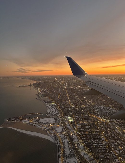 An aerial view from an airplane window at sunset, showing the wing over a snow-covered coastal city with glowing streetlights.