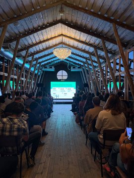 An audience sits in a rustic wooden venue with a chandelier and exposed beam ceiling, attentively watching a presentation displayed on a large screen at the front. The gathering reflects a Solana-focused event in Georgia, bringing together local builders and the Superteam community to discuss and engage with the ecosystem.