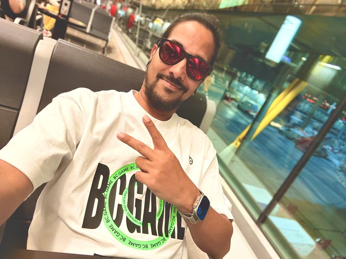 A bearded man wearing red-tinted sunglasses and a white BC GAME t-shirt flashes a peace sign while taking a selfie in what appears to be an airport terminal. The casual, friendly pose perfectly matches the laid-back "GM fam" (good morning family) greeting from his social media post.