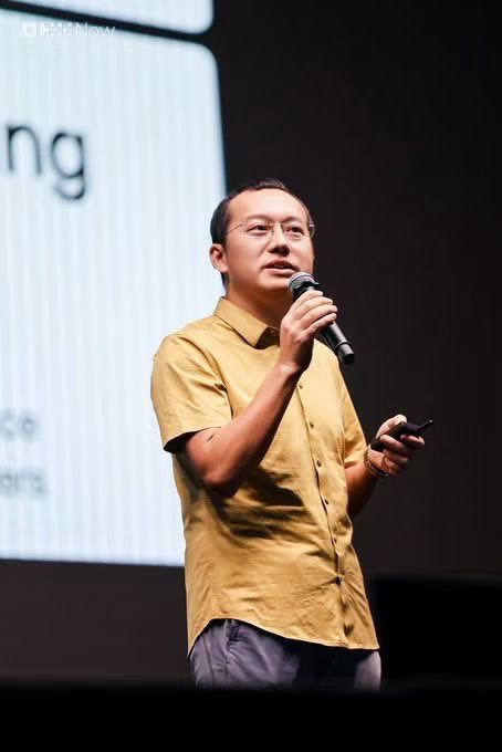 A man wearing glasses and a mustard yellow short-sleeved shirt speaking into a microphone while holding a presentation clicker, appearing to give a talk on stage.