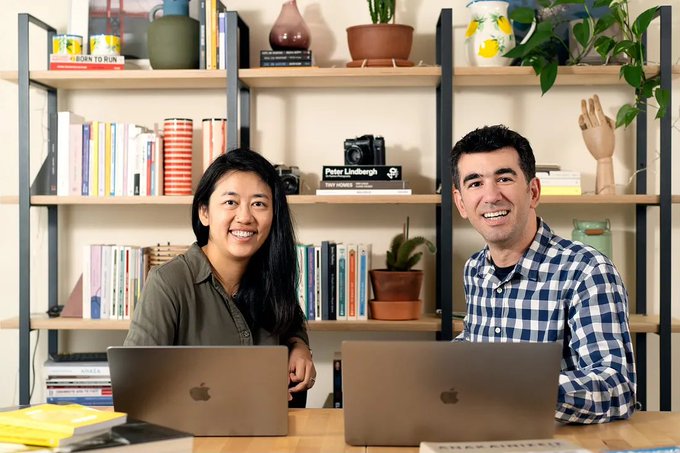 A woman with long dark hair and a man in a blue checkered shirt smile at the camera from behind two laptops, seated in front of a bookshelf displaying books, a camera, and a "Peter Lindbergh" book. The image illustrates the accompanying post's reference to Chengdiao Fan and Nicolas Kokkalis, Stanford PhDs who developed the Pi Network with a vision of fair crypto distribution.