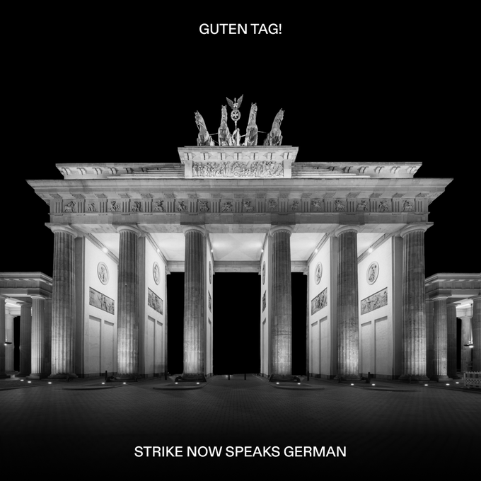 A black-and-white nighttime photo of the illuminated Brandenburg Gate in Berlin with the text "GUTEN TAG!" at the top and "STRIKE NOW SPEAKS GERMAN" at the bottom, visually celebrating Strike's expansion of language support to include German.