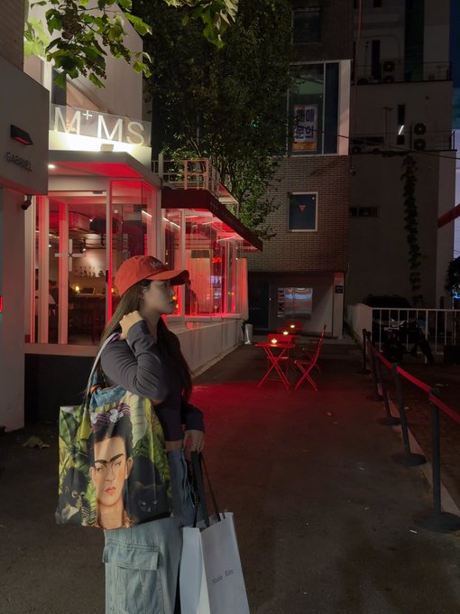A young woman wearing a red cap and a tote bag featuring a portrait stands outside a building with red lighting and signage reading 'M+MS' and 'GABRIEL'.