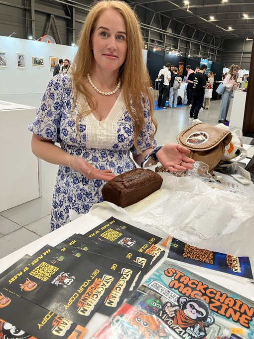 A woman with long red hair and a pearl necklace holds up a loaf of banana bread at BTC Prague, with "STACKCHAIN" and "Stackchain Magazine" promotional materials visible on the table in front of her.