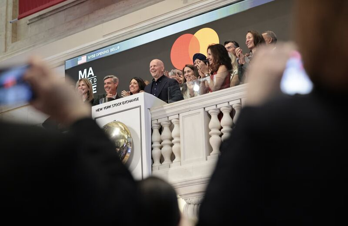A group of people applaud at the New York Stock Exchange podium during the Mastercard (MA) IPO opening bell ceremony, viewed from behind spectators taking photos.