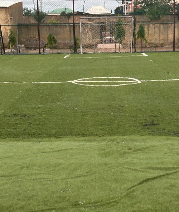 An empty soccer field with a goal in the background, enclosed by a chain-link fence with trees and a building visible behind it.