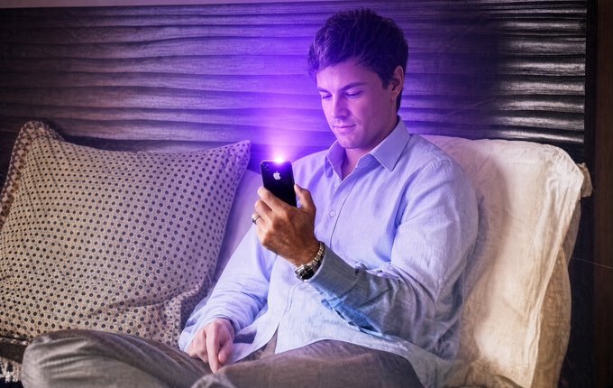 A man in a blue shirt sits on a couch at night, illuminated by the glow of his smartphone, which has a small purple light attachment on the back.