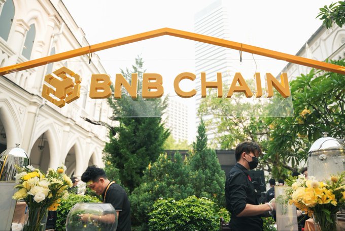 Staff members prepare a food display beneath a prominent orange "BNB CHAIN" sign at an event, likely tied to the #BNBSG social media campaign referenced in the post. The BNB Chain branding and elegant venue decor suggest a promotional gathering or community event celebrating the blockchain network.