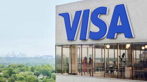 An image of a modern building with a large blue 'VISA' logo on its exterior, showing people inside through the windows and a cityscape in the background.