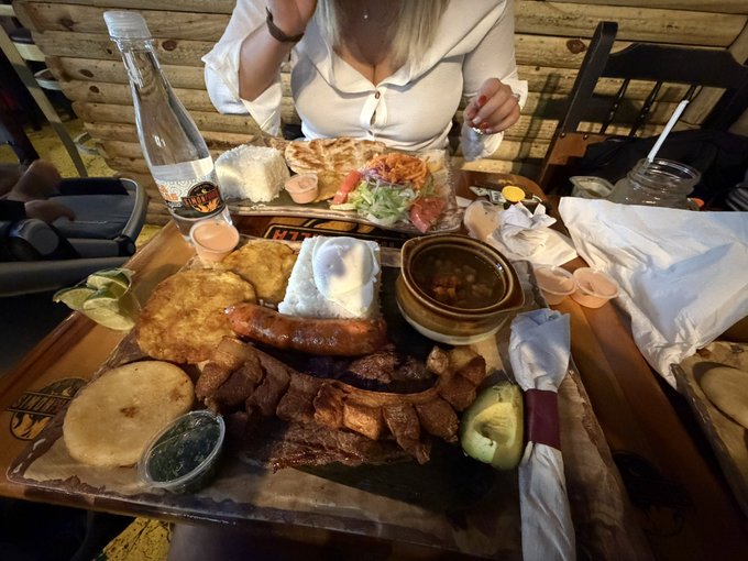 A person sits at a restaurant table in front of a large wooden platter piled high with a hearty meal of meat, potatoes, rice, and various sides.