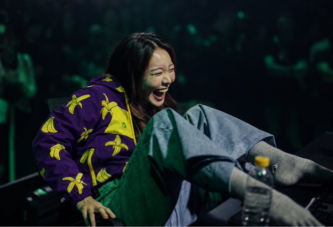 A laughing woman wearing a purple jacket with yellow banana prints sits in front of an audience, with a water bottle visible nearby.