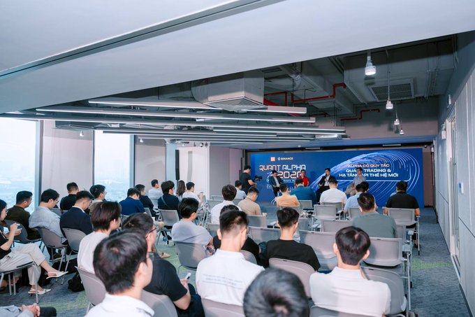 An audience attends a 'QUINT ALPHA 2026' Binance seminar event focused on trading, with attendees seated facing a stage featuring speakers and a large presentation screen.