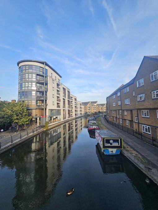 A serene view down a canal lined with brick apartment buildings and a modern glass-fronted structure, with narrowboats moored along the water and a duck swimming in the foreground under a blue sky with wispy clouds.