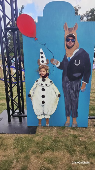 A man and a young girl pose at an outdoor event in costumes standing in front of a blue backdrop; the man is dressed in a donkey costume and the girl in a clown costume, with 'ClickOnChain' watermarked in the corner.