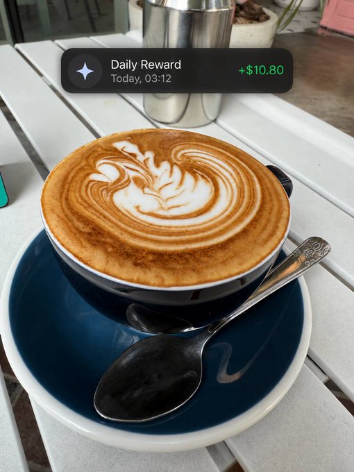 A latte art in a blue-rimmed cup sits on a slatted outdoor table beneath a digital "Daily Reward +$10.80" notification dated Today, 03:12—visually reinforcing the caption's message that Ethena Pay turns everyday moments (like grabbing a coffee) into automatic earnings, making rewards the default state of money in the account.