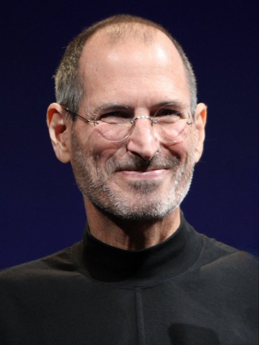 Portrait of Steve Jobs smiling slightly, wearing his signature round wire-rimmed glasses and a black mock turtleneck against a blue backdrop.