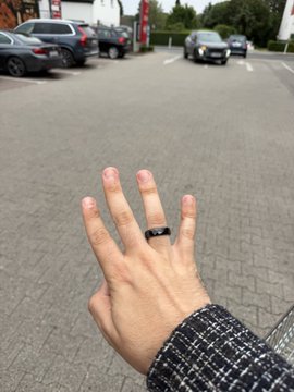 A hand wearing a sleek black Tangem Ring is raised against a backdrop of a parking lot with parked cars, showcasing the ring's minimalist design in everyday life. The image complements the post's theme of the ring seamlessly integrating into a stylish lifestyle, paired here with a tweed-patterned sleeve that highlights the ring's modern, adaptable aesthetic.