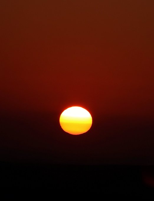 A vibrant sunset with the sun appearing as a glowing orange orb low on a dark horizon against a deep red sky.
