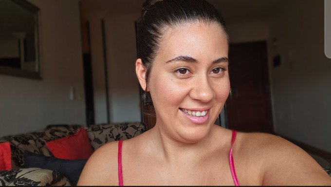A smiling woman with dark hair pulled back, wearing a pink top, sitting on a patterned couch indoors.