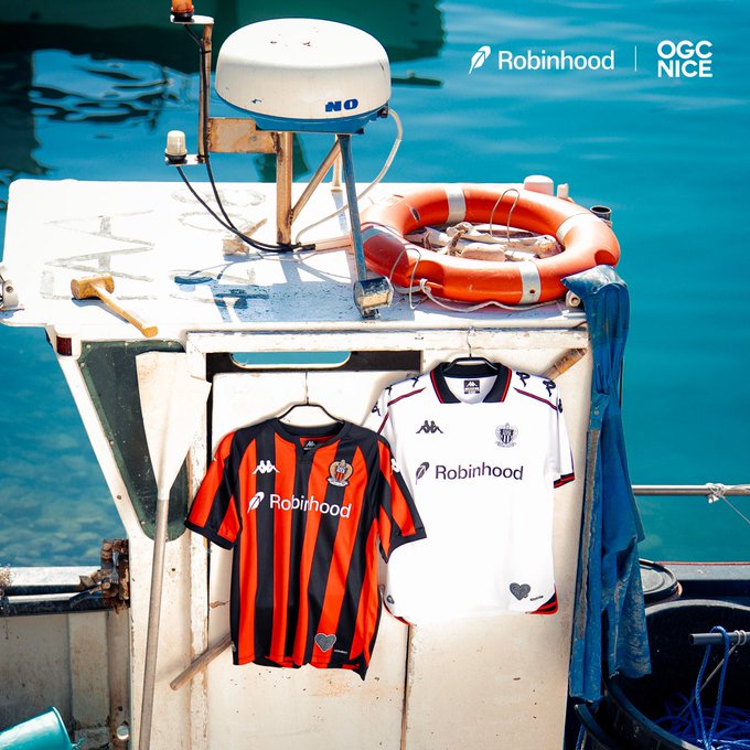 Two soccer jerseys hang on a boat against the sea, featuring Robinhood and OGC Nice logos, with the orange and black striped jersey on the left and the white jersey on the right.