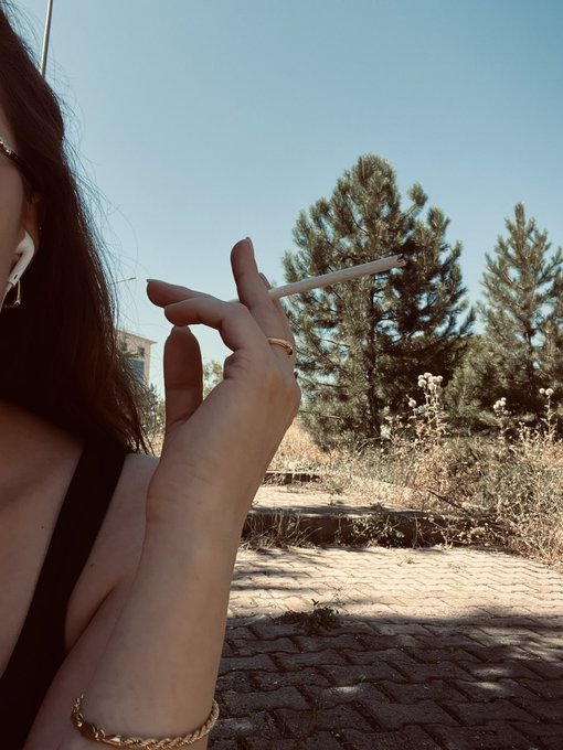 A woman with long dark hair and an airpod holding a lit cigarette, photographed outdoors on a stone path with pine trees in the background.