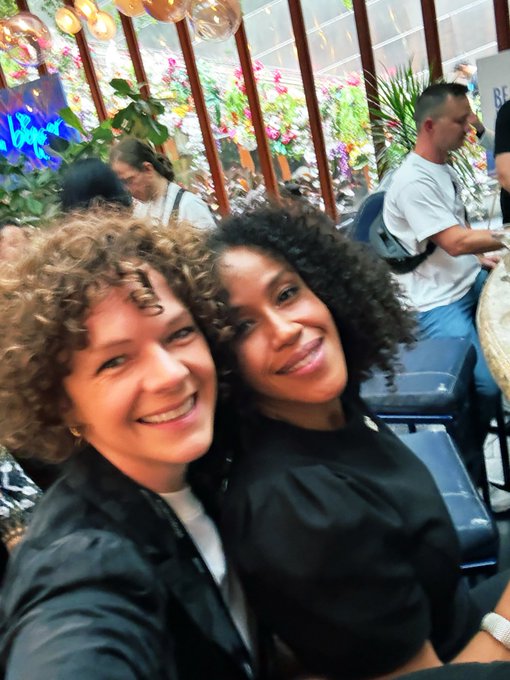 A selfie of two smiling women with curly hair posing closely together at what appears to be a social gathering or event, with a man seated in the background among plants and decorative lights.