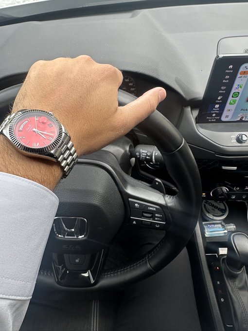 A close-up photo of a person's hand on the steering wheel of a Honda car, wearing a silver Rolex Datejust watch with a red dial, showing the dashboard infotainment screen and a Yankee air freshener.