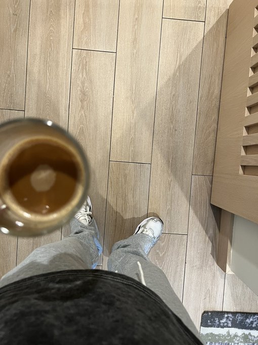 A first-person view looking down at sneakers and jeans on wood flooring, with a glass of coffee held in the foreground—the perfect morning scene to match a cheerful "Gm" Sunday greeting.