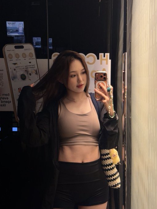 A young woman takes a mirror selfie wearing a fitted tank top, black shorts, and an open jacket, looking energetic and ready to start her day. Partially visible posters in the background include Apple's "Shot on iPhone" campaign ("Shot with iPhone 15 Pro Max," "A camera that captures light, shadow, color, and detail"), tying into her 7 a.m. class to 8 a.m. routine as a "high-energy person."