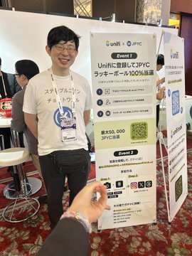 A man wearing glasses and a graphic t-shirt standing next to a Unifi + JPYC promotional banner advertising 'Unificに登録してJPYCラッキーボード100%当選' with a 50,000 JPYC reward and event 2 details.