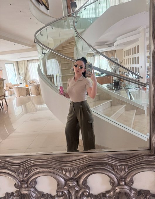 A mirror selfie of a woman wearing sunglasses, a beige polo shirt, and olive green cargo pants, standing in front of a curved staircase in a luxurious home.