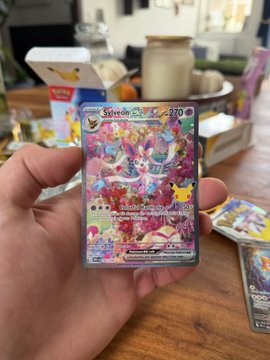 A hand holds up a holographic Pokémon trading card featuring Sylveon EX with 270 HP, set against a background of other cards and items on a wooden surface.