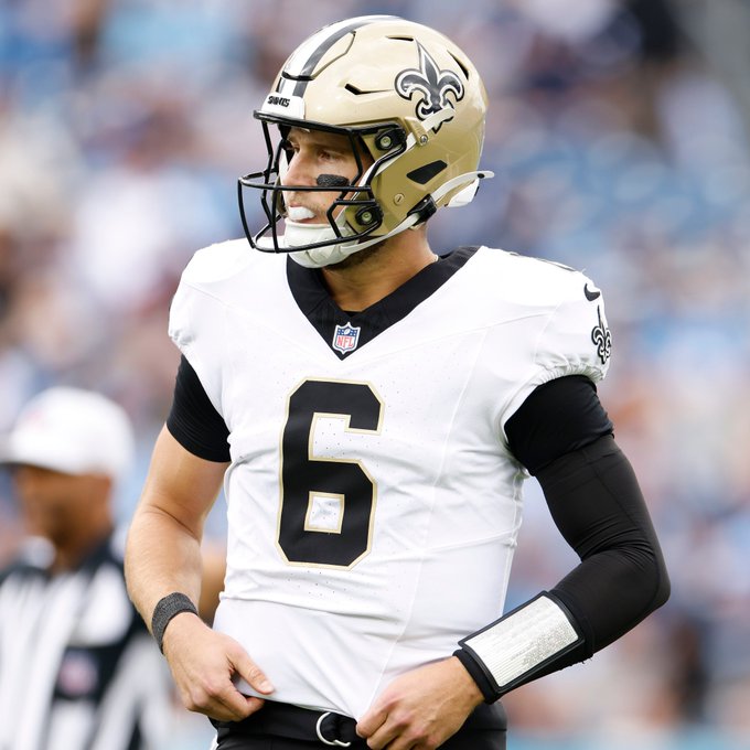 New Orleans Saints quarterback #6 (Spencer Rattler) stands on the field wearing a white jersey with a gold helmet, appearing disappointed during a game against an opposing team.