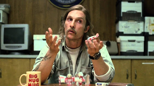 A man with long hair and a mustache in a green shirt gestures with his hands while sitting at a desk, with a 'BIG HUG MUG' mug and small figurines visible in the foreground.