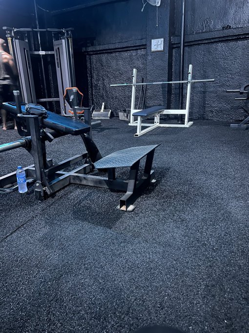 An empty home gym with black rubber flooring, featuring weight benches, a squat rack, and cable machines against dark textured walls.