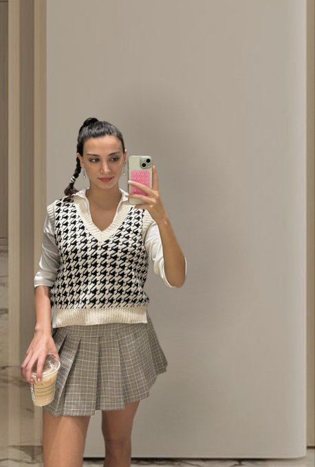 A mirror selfie of a young woman wearing a houndstooth sweater vest and plaid skirt, holding a pink phone case and a coffee cup.