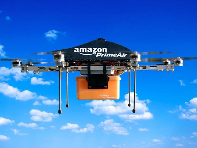 An Amazon Prime Air delivery drone, prominently displaying the "amazon PrimeAir" logo, hovers in a bright blue sky while carrying an orange Amazon-branded package. The image visually reinforces the news of Amazon's expansion into drone delivery across hundreds of US cities, complementing the announcement about the tokenized $AMZN on the Solana blockchain.