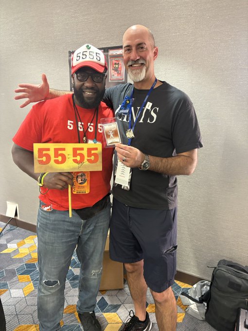 Two men pose together indoors, one wearing a red shirt and cap both featuring "5555," holding a matching sign, while the other wears a dark grey Giants t-shirt and holds a small award.